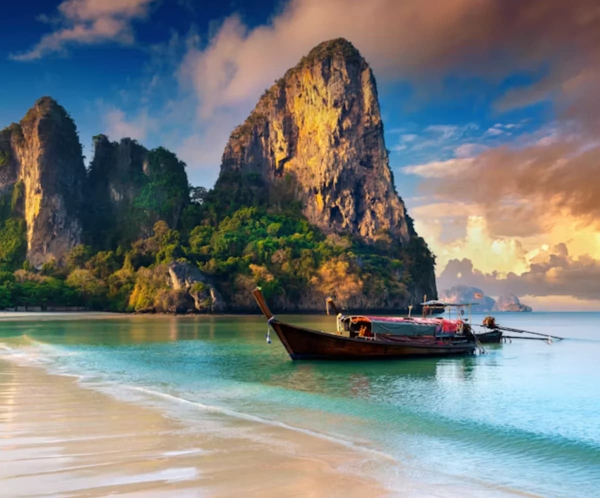 Long-tail boats moored in shallow turquoise water off a white-sand beach, beneath limestone karst cliffs on the Andaman coast