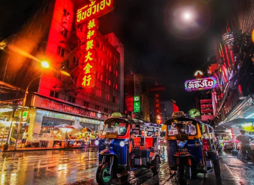 A Bangkok street in the evening, red neon signage glowing above tuk-tuks on wet tarmac