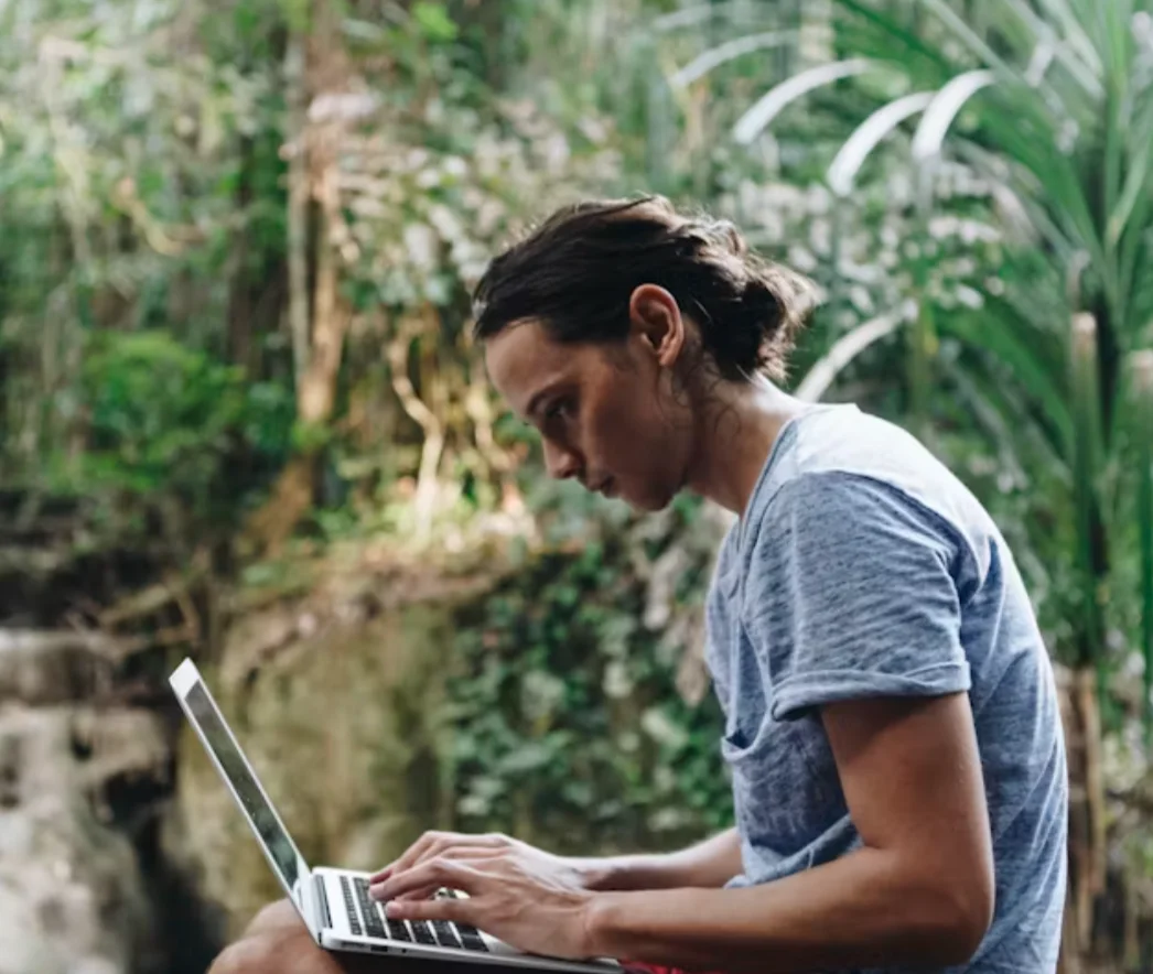 A person typing on a laptop balanced on their lap, sitting outdoors among dense tropical greenery